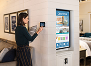 A woman adjusts a small digital wall panel in a retail store. A large digital display is nearby.