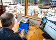 Over the shoulder of man at a laptop in an office overlooking a warehouse floor