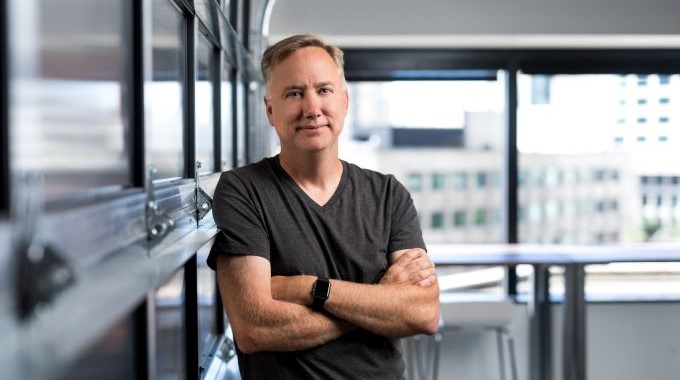 Portrait of man standing with arms folded and facing camera. He is wearing a grey tee shirt and smartwatch, leaning against a closed garage door in an indoor space with windows in the background