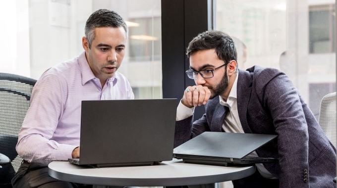 Two men sitting at a table looking at one of their laptop screens in discussion